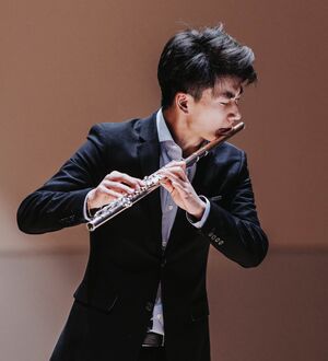 Person wearing a suit playing the flute on stage, focused on their performance, with a poised and professional presence.