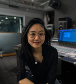 Person wearing a black button-down shirt sitting on a stool in the electronic music studio, surrounded by equipment and sound gear.