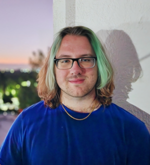 Headshot of a person with shoulder-length hair featuring green streaks in the front, wearing a blue shirt, standing in front of a wall.