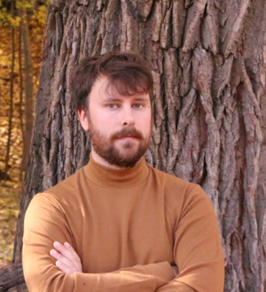 Headshot of a person with a beard, wearing a brown turtleneck, standing in front of a tree in the woods.