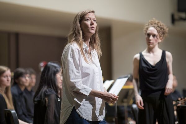 Person in a white shirt standing on stage, surrounded by an ensemble in performance attire, speaking to the audience, creating a connection before or during the performance.