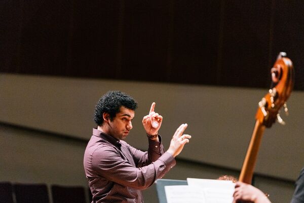 Conductor wearing a burgundy shirt leading an ensemble, with expressive hand movements and focused attention on the musicians.