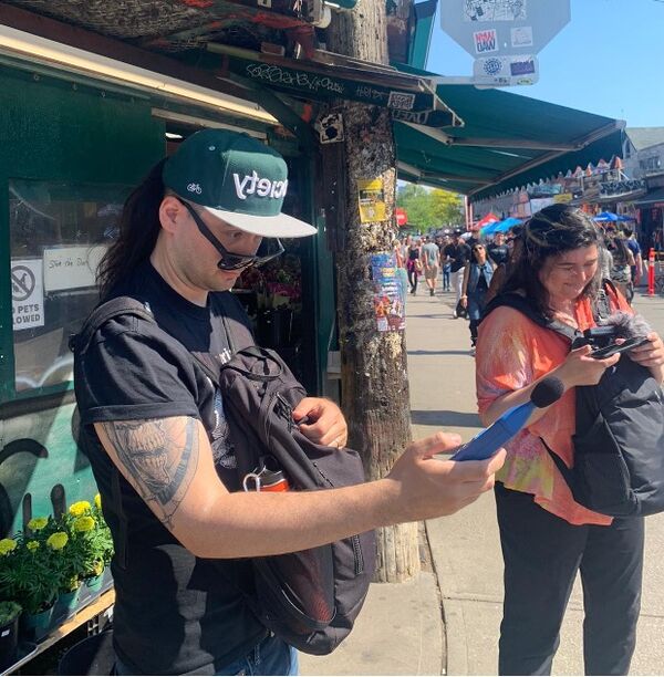 People wearing backpacks on their fronts, looking into the bags while standing at a market, engaging with and interviewing attendees in a lively setting.