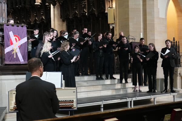 Choir in performance attire singing on risers on stage, with an accompanist playing a harpsichord.