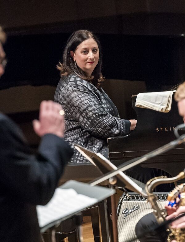 Woman in a grey jacket playing piano for an ensemble, focused on the conductor as she performs.
