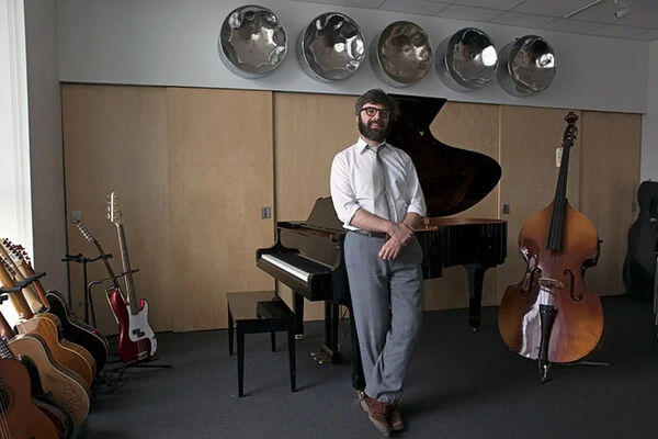 Man with a beard wearing business attire, standing in a music room surrounded by various instruments, including guitars, grand piano, and a double bass. 