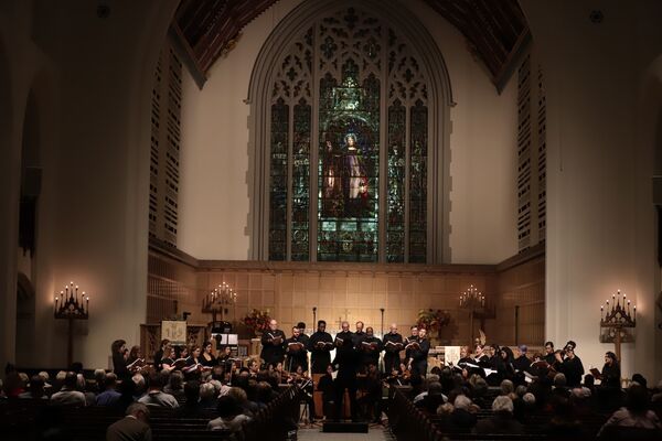 Choir performing in a church for an audience, with a stained glass window in the background.