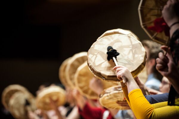 Group of people playing Indigenous-style drums, with the focus on a person wearing a yellow shirt striking a drum.