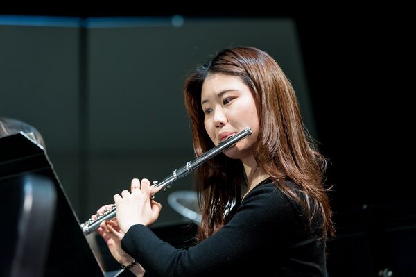 Person wearing performance attire playing the flute, focused on a music stand with sheet music, immersed in the performance.