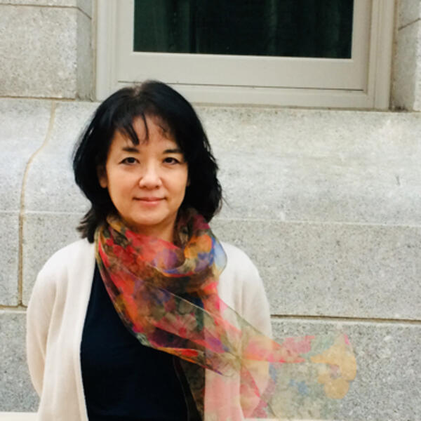 Person wearing a colorful scarf and white cardigan, standing in front of a stone building with a window.