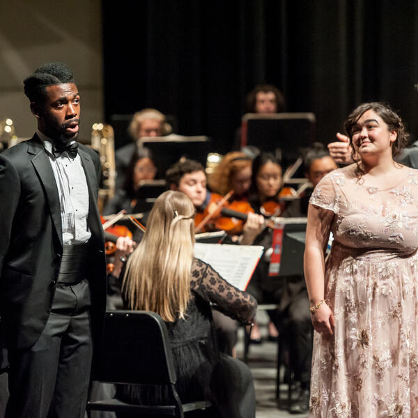 A male and female opera soloists singing in front of an orchestra